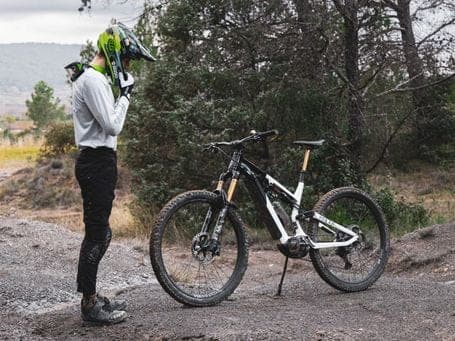 Road cyclist in fitted gear adjusting their neon yellow-green helmet next to a black and white mountain bike on a wooded mountain trail