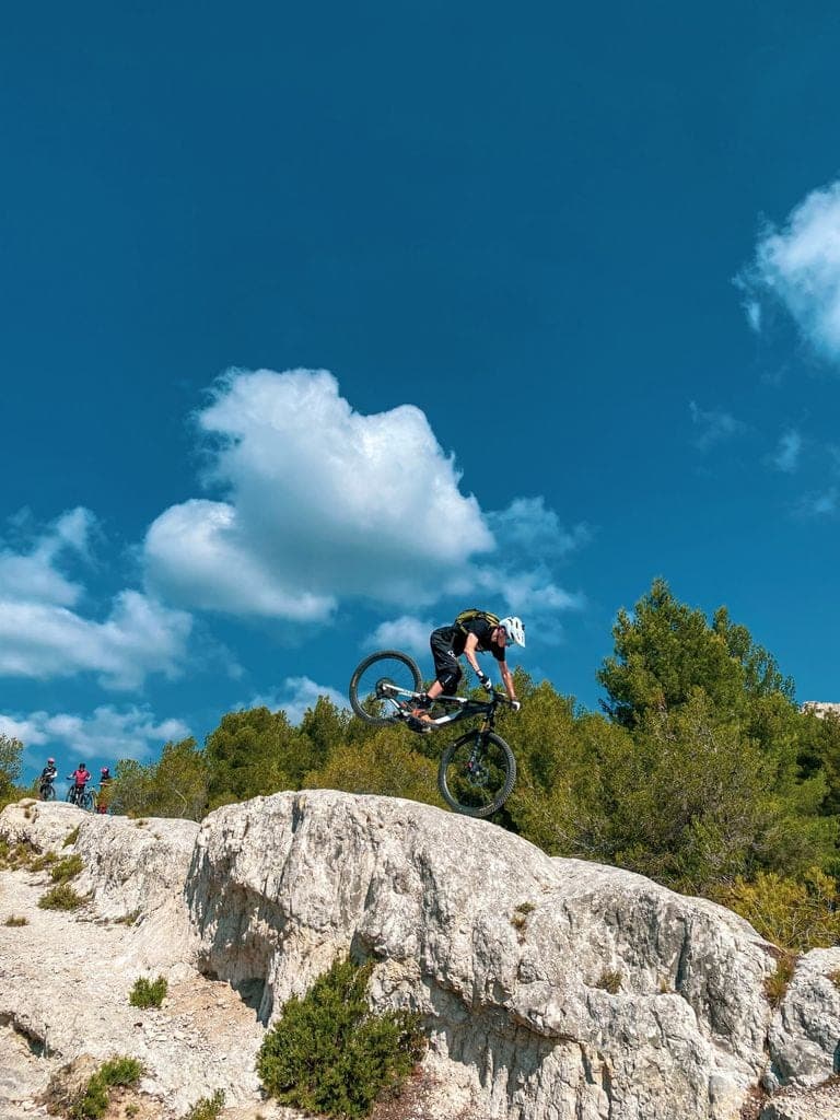 An MTB rider performs an acrobatic jump over white limestone rocks under clear blue sky, with spectators and Mediterranean vegetation in the background