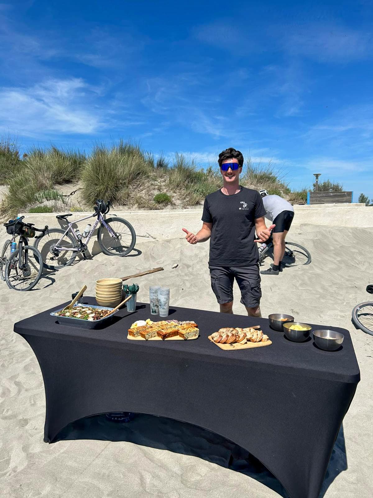 A man in cycling gear poses in front of a picnic table laden with food and drinks on a sandy beach, with bicycles and grassy dunes in the background on a sunny day.