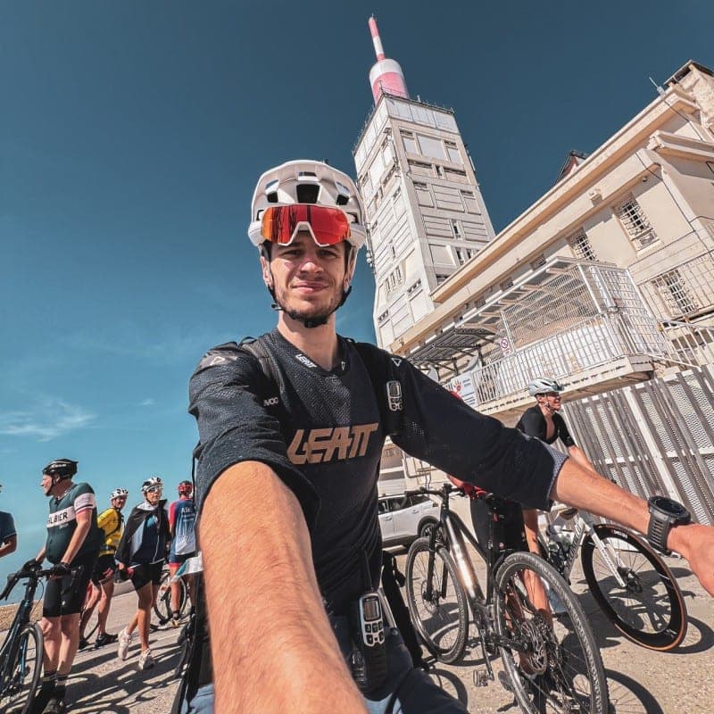 Cyclist in sports attire smiling at the camera with their bike in front of a historic tower in Provence on a beautiful day