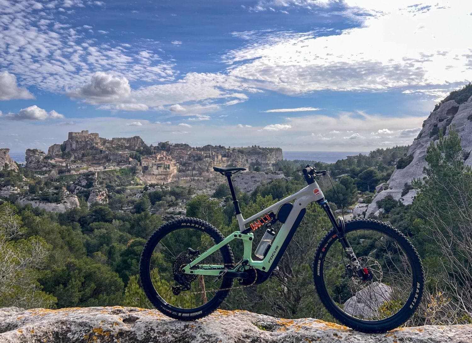 A green and white electric mountain bike resting on a rock, with a hilltop village perched on a rocky cliff in the background surrounded by forests and limestone formations typical of Provence