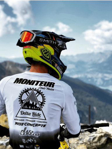 Mountain biker viewed from behind wearing a yellow and black Smith helmet and a Montreur t-shirt, facing the snow-capped Alps mountains