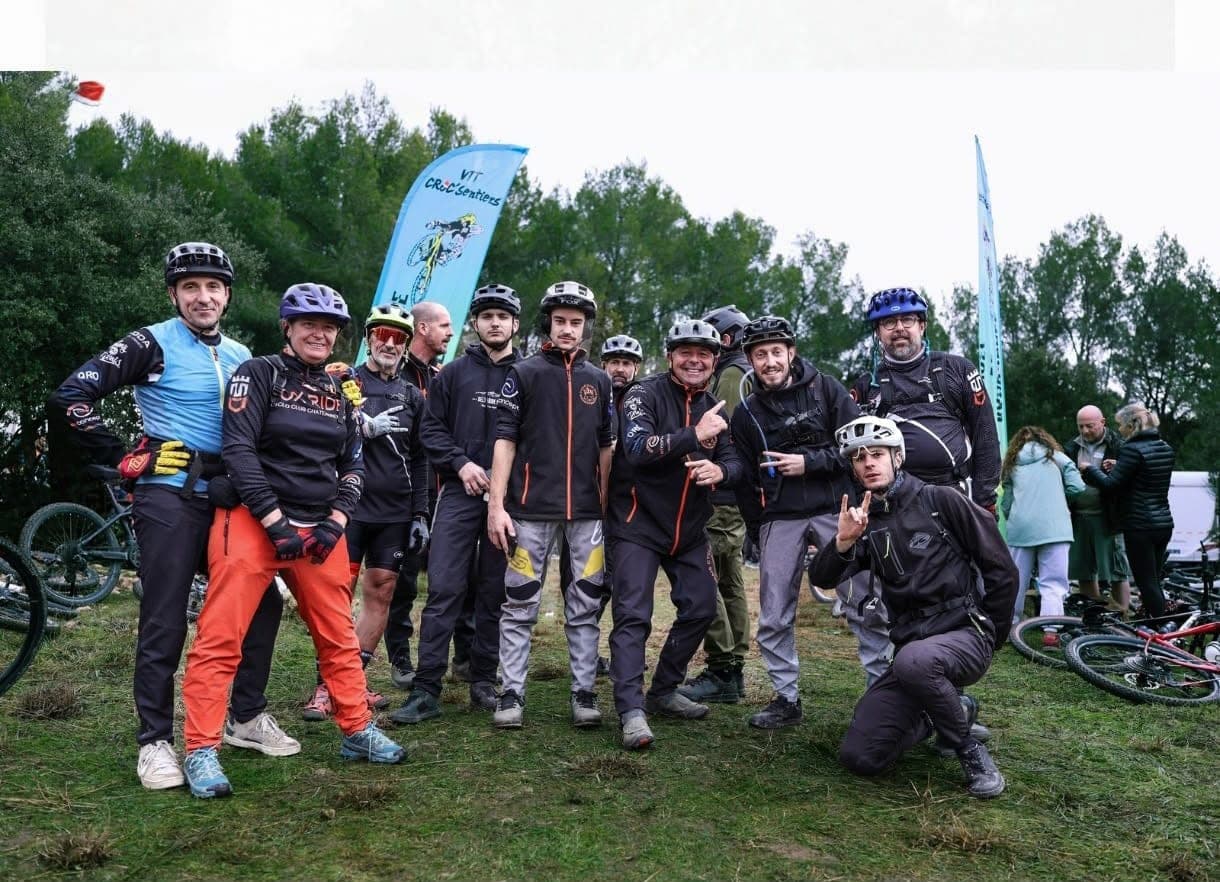 Group of smiling cyclists in electric mountain bike gear posing together in a field with blue sails and forests in the background during an outdoor event