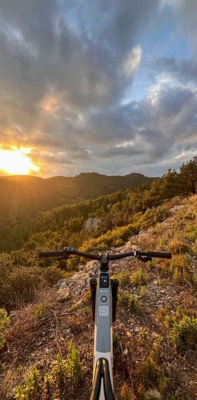 First-person view of an electric bike on a rocky mountain trail with sunset over wooded hills
